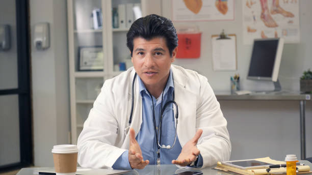 Male doctor speaks to camera during web conference A serious mid adult male doctor sits at his office desk and gestures as he speaks to the camera during a web conference. doctor utensils stock pictures, royalty-free photos & images