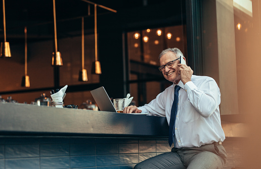 Smiling senior man in formal wear sitting at bar counter with laptop talking on cell phone. Happy mature entrepreneur making a phone call from bar counter.