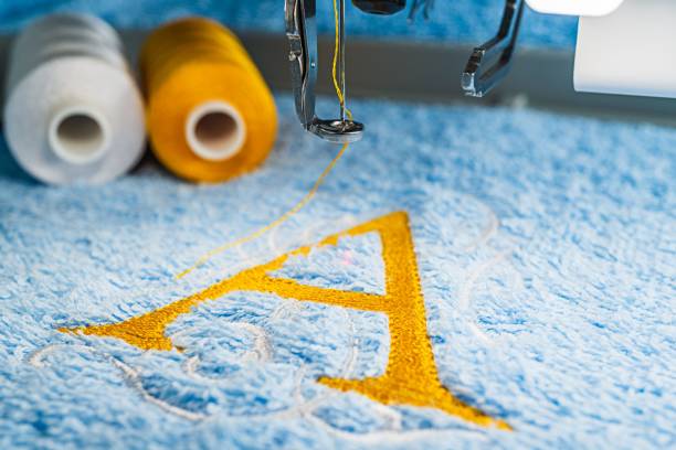 A alphabet design on towel in hoop of embroidery machine Yellow A alphabet logo design and two color threads on blue towel in hoop of embroidery machine, close up picture embroidery photos stock pictures, royalty-free photos & images