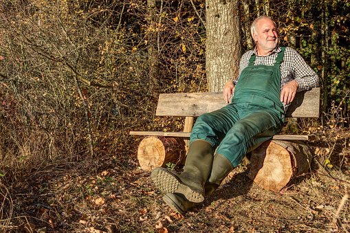 A farmerwith green dungarees and rubber boots relaxes after work on his bench at the edge of the forest in the mild evening sun.\