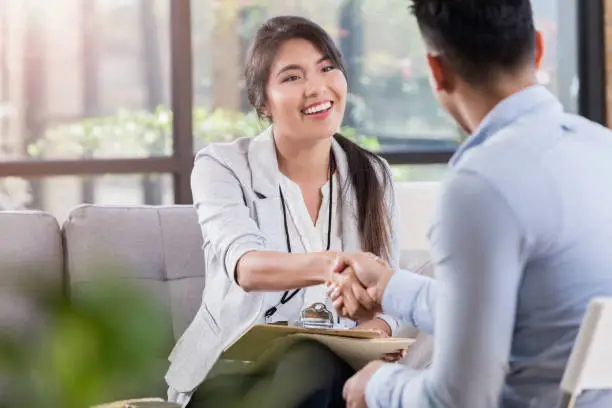 During an introduction before a counseling meeting, female doctor and male patient shake hands. Doctor sits on the sofa in her office while holding a clipboard and wearing a lanyard.