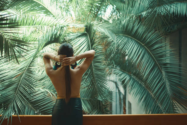 Back view fit exotic woman, bare back, watching huge palm tree leaves while resting on the balcony, arranging her hair, extending her hands up. Resort and vacations promotion. Beautiful life concept.
