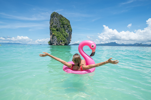 Young woman arms outstretched on idyllic beach with inflatable flamingo playing in pristine clear water in the Islands of Thailand. People travel luxury fun and cool attitude concept