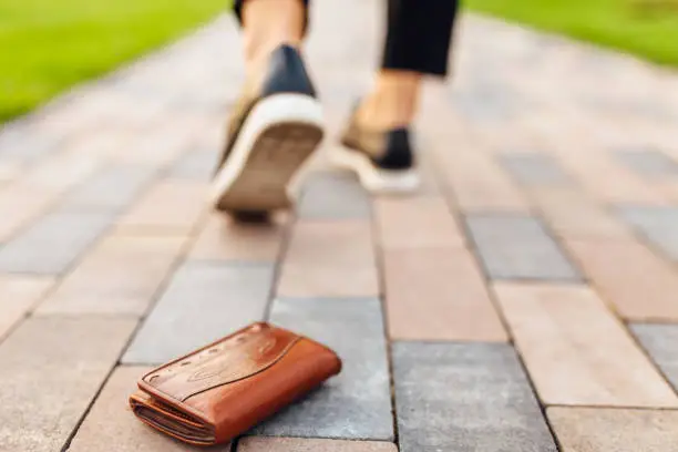 Photo of The girl lost a leather wallet with money on the street. Close-up of a wallet lying on the sidewalk and legs of a departing girl