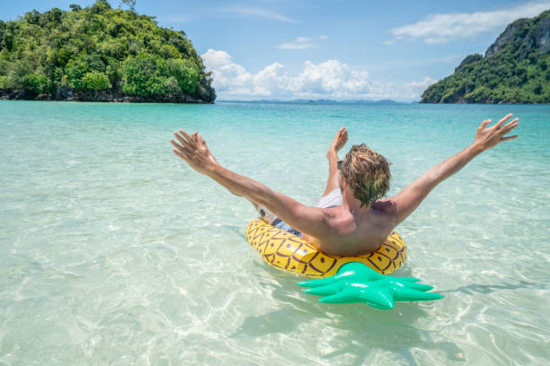 Joyful young man floating with inflatable pineapple on beautiful tropical beach enjoying beach holidays in paradise. Male outstretches arms for happiness and vitality