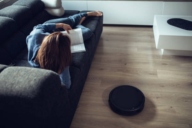 istock Woman reading book while robotic vacuum cleaner cleaning home 1058883946