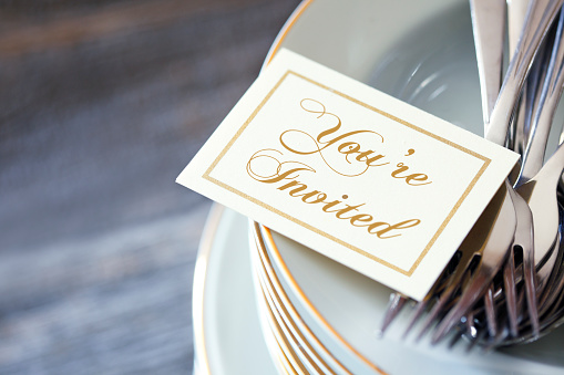 An invitation and a grouping of silverware rest on top of a stack of dinner plates. The image is photographed using a very shallow depth of field.