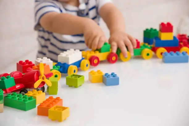 Photo of Close up of child's hands playing with colorful plastic bricks at the table. Toddler having fun and building out of bright constructor bricks. Early learning.  stripe background. Developing toys