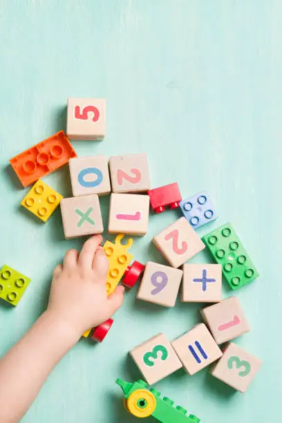 Photo of Child playing with wooden cubes with numbers and colorful toy bricks on a turquoise wooden background. Toddler learning numbers. Hand of a child taking toys.