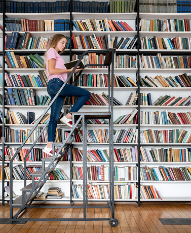 Female student looking for a book at the library climbing on a ladder â education concepts