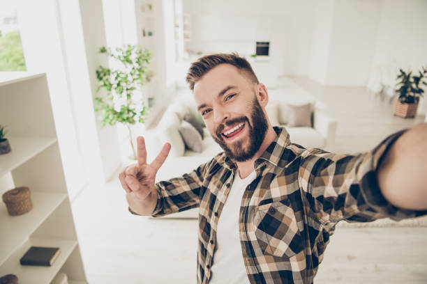 istock Fooling around and grimacing handsome man make selfie on the front camera of smartphone stand in checkered shirt and show v-sign 1059056994