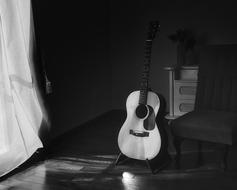 Black & white photo of acoustic Spanish guitar on a stand in the moody shadows of a dark room with bright light coming in from behind a curtain