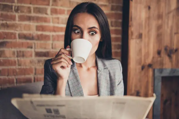 Photo of WOW! Close up portrait of astonishment brunette woman with crazy emotion and stare open eyed read the news about high oil prices drink espresso beverage from a white small mug in loft workstation