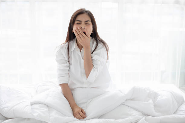 Asian woman Beautiful young smiling woman sitting on bed and stretching in the morning at bedroom after waking up in her bed fully rested and open the curtains in the morning to get fresh air. stock photo