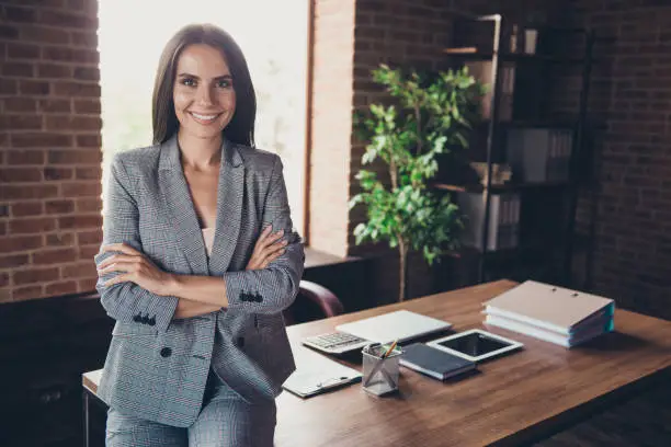 Photo of Beautiful elegant classic trendy latin smiling lady freelancer, company founder with folded crossed hands, wearing gray suit, invites to her team, at work place, station, modern fashionable loft