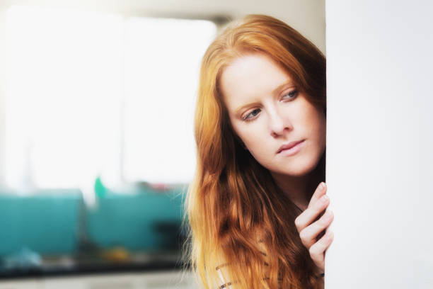 Young woman peeps suspiciously round wall, checking for intruders A pretty young redhead peeps round a corner in her home, looking suspicious as she checks for intruders. looking around stock pictures, royalty-free photos & images