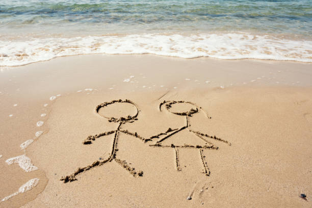 A beautiful shot of two stick figures holding hands drawn in sand with splashing waves on a hot summer day.