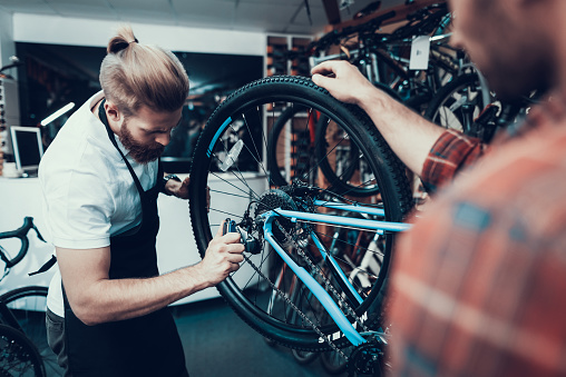 Professional Bike Repairman Fix Cycle in Workshop. Portrait of Young Caucasian Man Wearing Apron Examines Modern Cycle Transmission System Holds Hex Wrench. Bike Maintenance and Sport Shop Concept