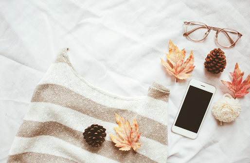 Autumn fashion style concept, sweater and smartphone with maple leaves on white bed sheet background