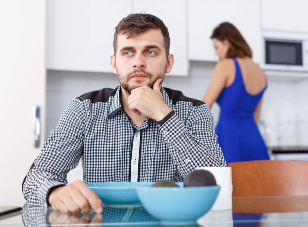 istock Young male sitting at kitchen table while woman preparing food 1059142558