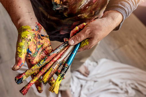 Photo of Unrecognizable male Artist holding paintbrushes in his art studio during the day. Closeup shot of paintbrushes and hands stained with paint. Cropped shot of male artist hands holding paintbrushes. Dirty hands holding brushes.