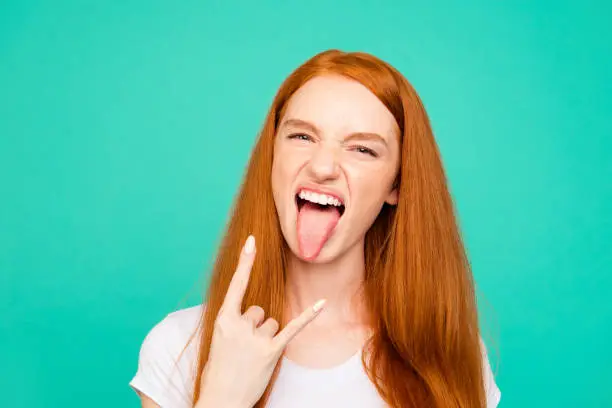 Portrait of nice positive cheerful foolish cute bright vivid shiny red straight-haired girl in casual white t-shirt, showing tongue out and rock-n-roll sign, isolated over turquoise green background