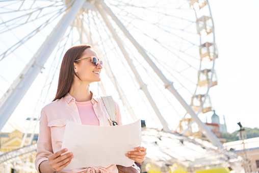 smiling attractive tourist in sunglasses standing with map near observation wheel and looking away