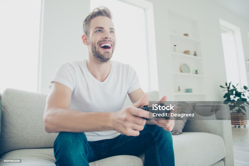 Low angle top view of handsome, attractive, sporty brunet bristle man in white t-shirt and blue jeans sit in light modern interior play at video games and rejoice about win Addiction Stock Photo