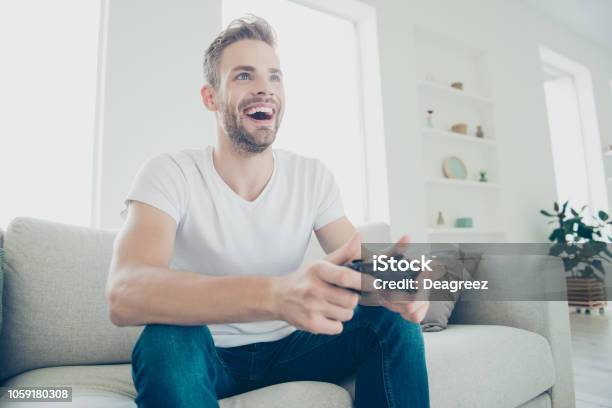 Low Angle Top View Of Handsome Attractive Sporty Brunet Bristle Man In White Tshirt And Blue Jeans Sit In Light Modern Interior Play At Video Games And Rejoice About Win Stock Photo - Download Image Now