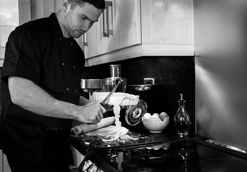Handsome male chef, dressed in black jacket, cuts off excess pastry from a dish