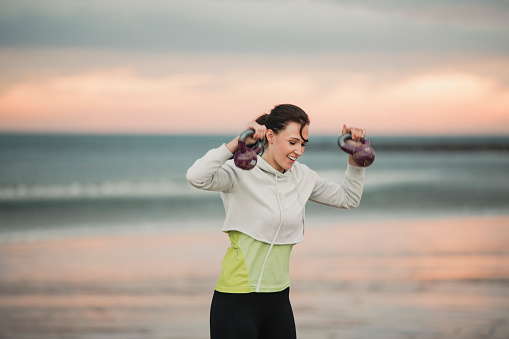 Portrait of a woman lifting kettlebells on a beach early hours in the morning. The sun is rising and is leaving a pink reflection on the sea.