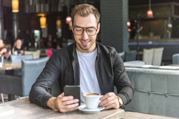 istock smiling young man with cup of coffee using smartphone in cafe 1059205252