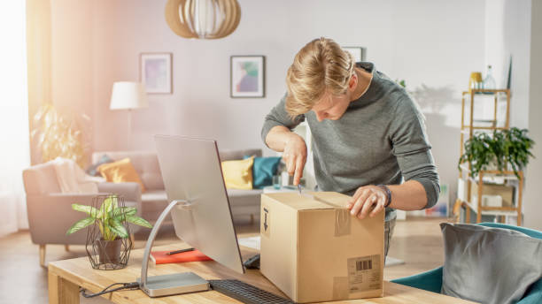 istock Handsome Young Man in Living Room with Cardboard Box Package, Opening it With Interest, Using Knife. 1059208276