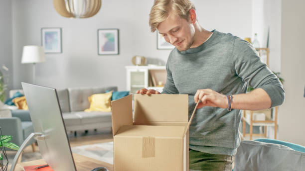 Handsome Young Man in Living Room Opening Cardboard Box Package With Interest.