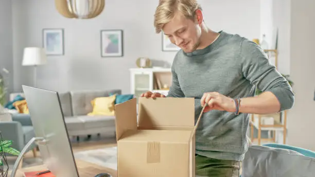 Photo of Handsome Young Man in Living Room Opening Cardboard Box Package With Interest.