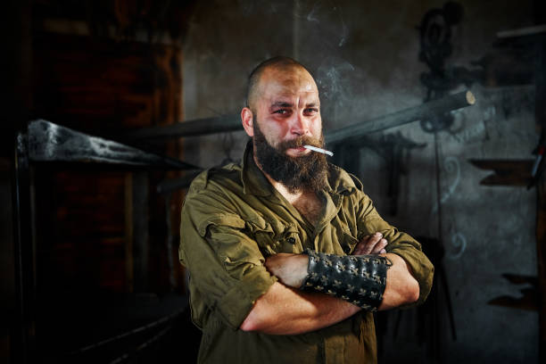 istock Brutal bearded blacksmith worker smokes after hard work. The man is tired, but smiling, his face is covered in mud and sweat 1059228502
