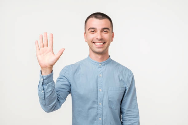 istock Attractive european student waives hand in hello gesture while smiling cheerfully over white background. 1059263134