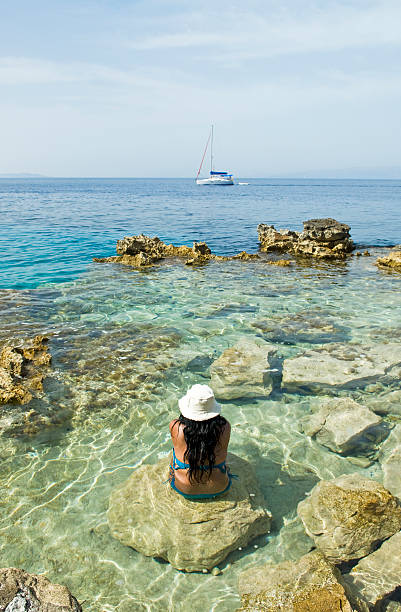 color portrait photo of a dark haired woman sitting on a rock and enjoying the view in the beautiful clear blue water while on her vacation to Greece.
