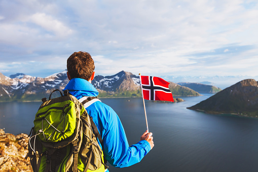 travel to Norway, hiker traveler with backpack holding norwegian flag and looking at beautiful fjord landscape