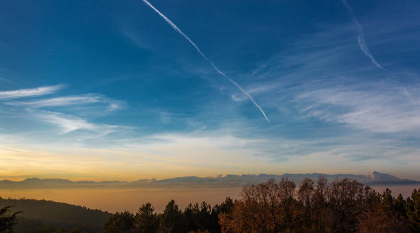 Landscape photography of wide blue sky in winter, with plane contrails, mountain ridge, fog covering the valley, trees. Sunset, sunrise, sparse clouds, steam from condensation. Photo taken in Macedonia.
