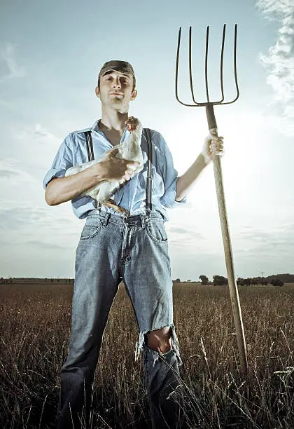 A young farmer standing in farm field with chicken and a pitchfork.