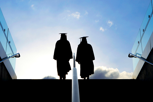 A shot of two college graduates as they make their way up a stairway and into the morning sunlight.  They are dressed in graduation robes and hats.  Lots of excitement and promise ahead of them.