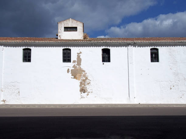 old white painted building with high small windows and a tiled roof alongside a black roof with bright sunlit cloudy blue sky an old long white painted building with high small windows and a tiled roof alongside a black roof with bright sunlit cloudy blue sky rood stock pictures, royalty-free photos & images