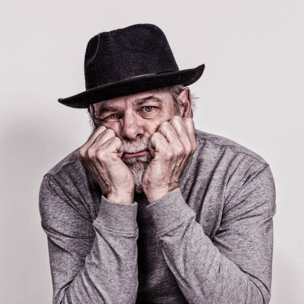 A sad looking, melancholic senior adult man looking at the camera with his head in his hands. He is wearing a shabby, grubby, black felt fedora hat covered with tangled cat hair, dust and random fuzz.