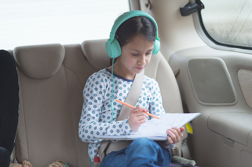 A young girl in the back seat if a car draws and colors in a book while she listens to music on over-the-ear headphones.