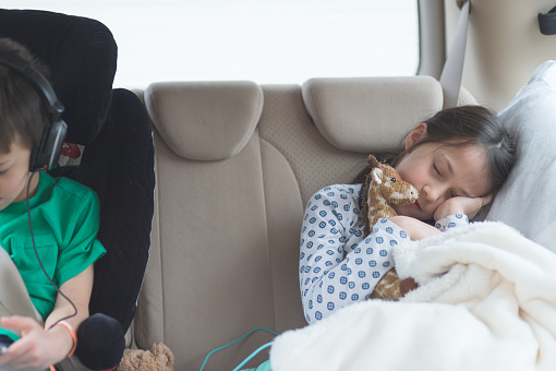 A young elementary-age girl takes a nap in the back seat of the car on a long road trip. She is snuggled up with a blanket and a stuffed animal giraffe. Her brother is in a booster seat next to her playing a game on a tablet.