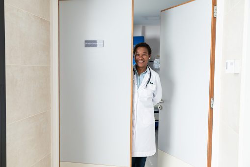 Friendly african american female doctor standing behind the door of her private practice smiling at camera very happy