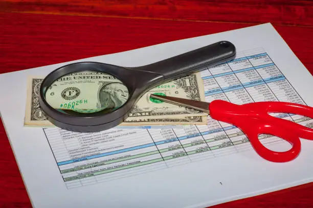 Budget tools - Instruction to the management team of the company to reduce business operational costs. The pair of scissors signifies the intention to cut operational costs (be wise); the magnifying glass, to look for savings and to find every dollar that can be saved (be innovative). The glass distorts the dollar. Spreadsheet prepared as a prop and has no real-life significance. Image shot in studio environment. Camera: Canon EOS 5D MII. Lens: Leica APO Summicron-R 90mm F2 ASPH.
