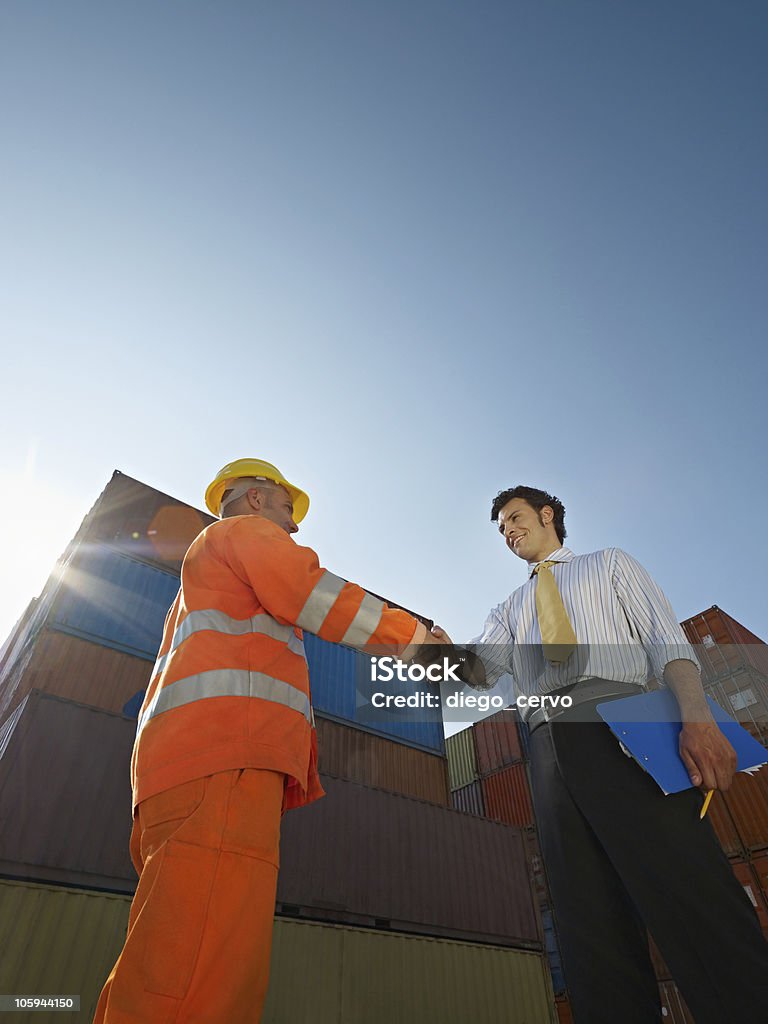 businessman and manual worker with cargo containers Mid adult businessman holding clipboard and shaking hands to manual worker near cargo containers. Vertical shape, low angle view. Copy space 30-39 Years Stock Photo