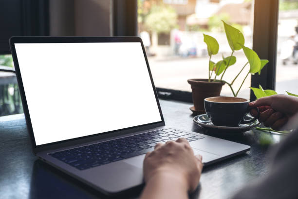 istock Mockup image of a woman's hands using and touching laptop touchpad with blank white desktop screen while drinking coffee on wooden table in cafe 1059442176
